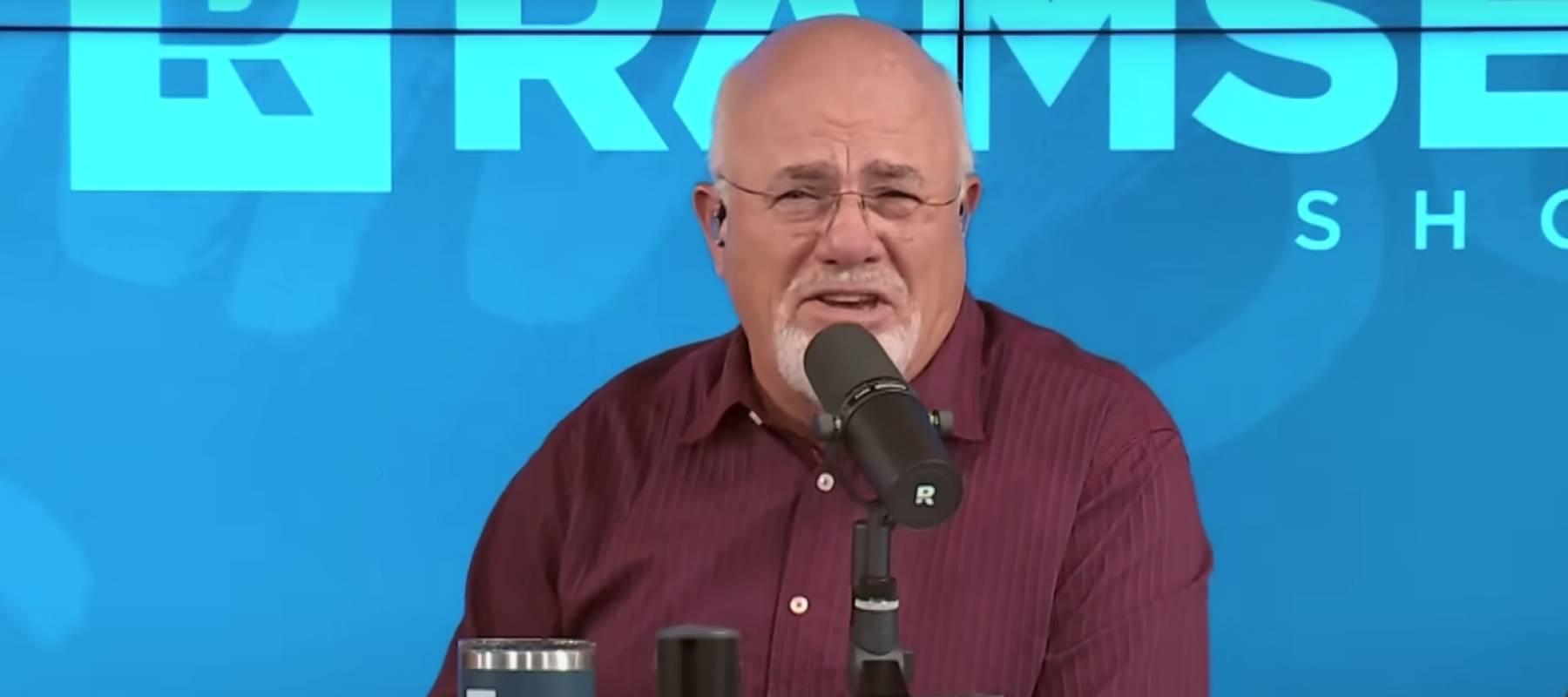 Dave Ramsey seen on set of his podcast, making an angry face and speaking into a microphone.
