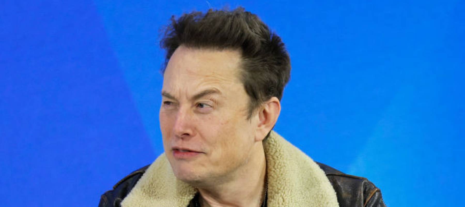 Elon Musk seen making a goofy face while on stage with a blue background.