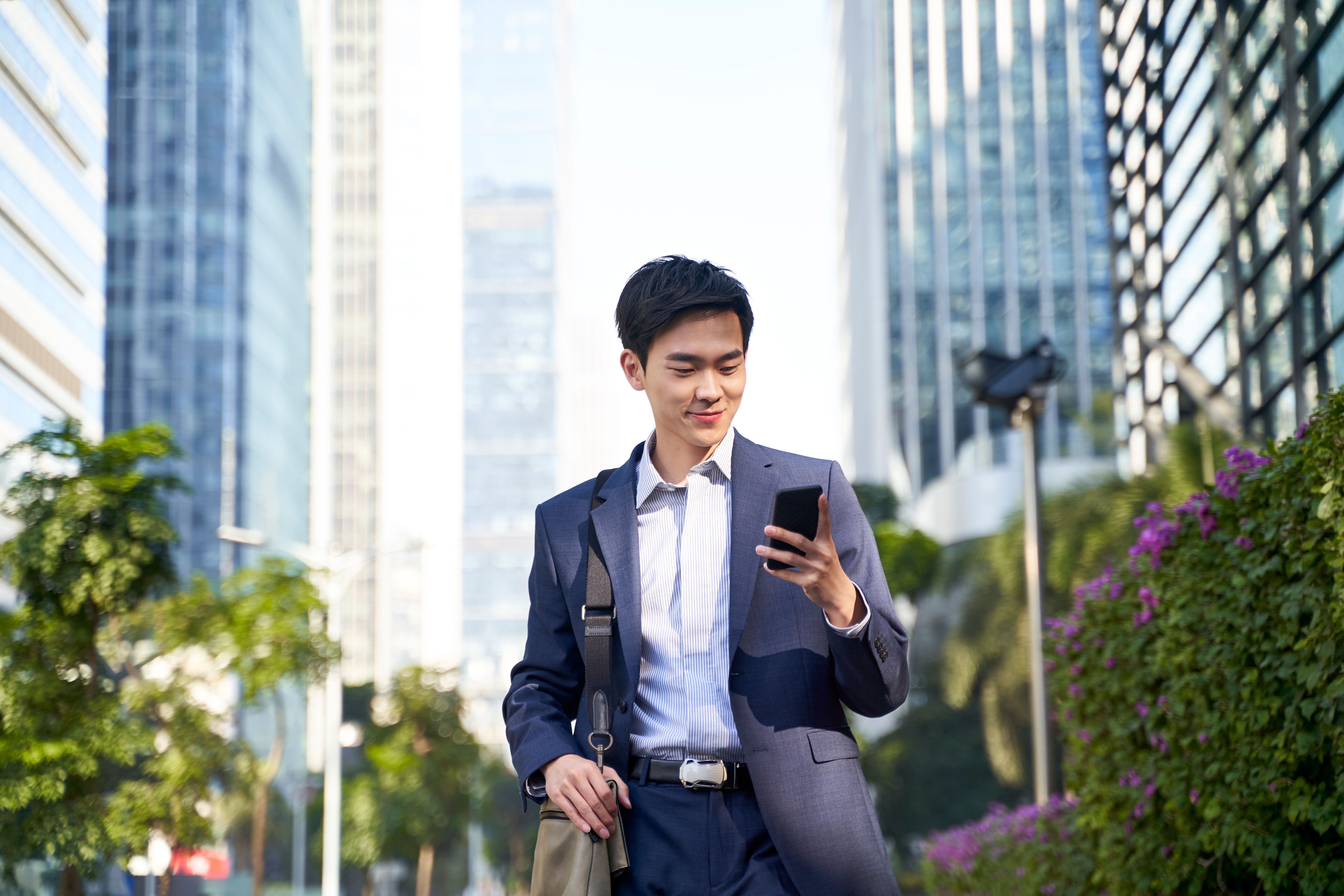 Young Asian businessman looking at messages on cellphone while walking in the street in downtown of modern city